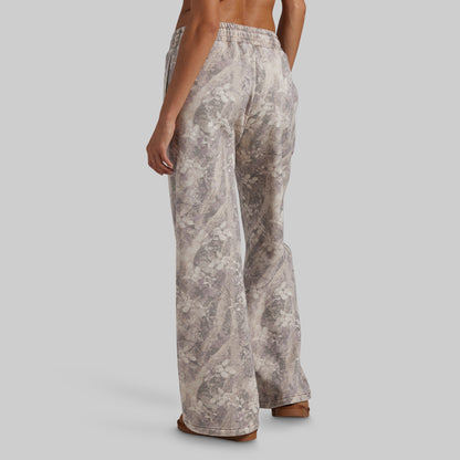 A person shown from the back wearing wide-leg pants with an abstract floral or marble-like print in beige, taupe, and gray tones, against a plain light gray studio background.