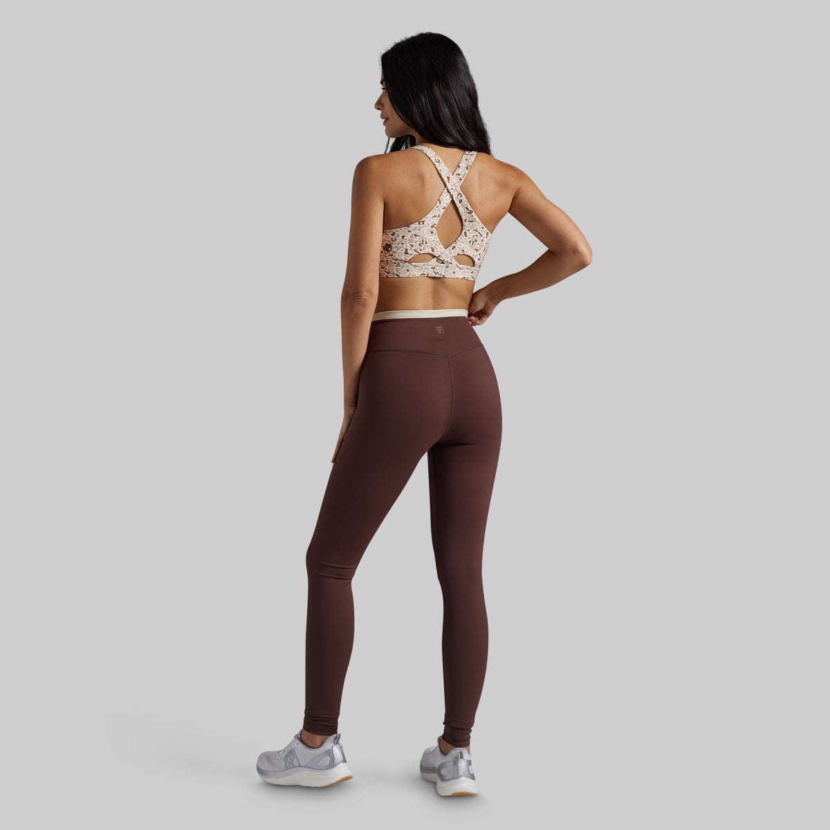 A woman is shown from the back wearing a brown athletic leggings set and a patterned beige and brown sports bra, standing against a solid gray studio background.