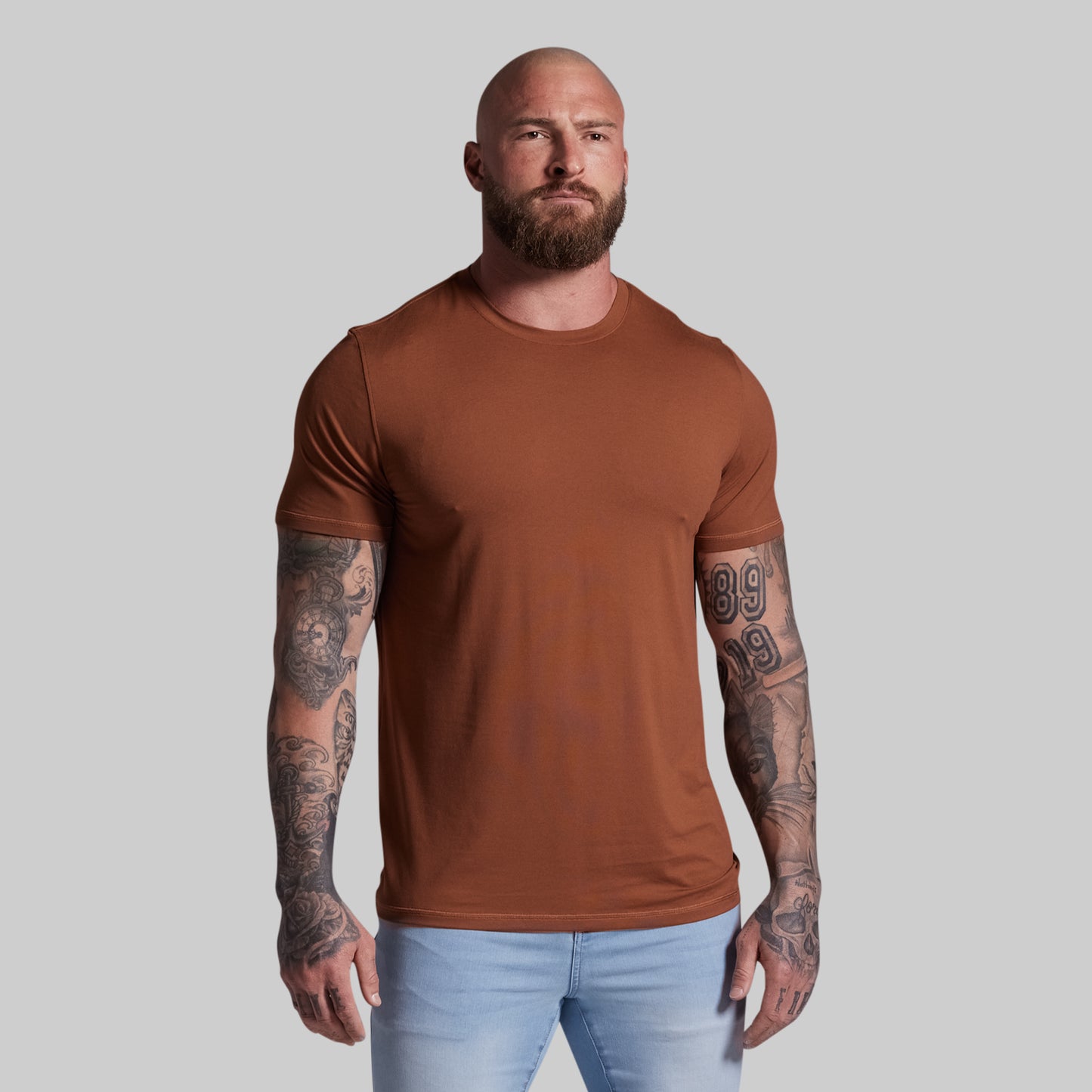 Man wearing a brown t-shirt and light blue jeans against a plain background