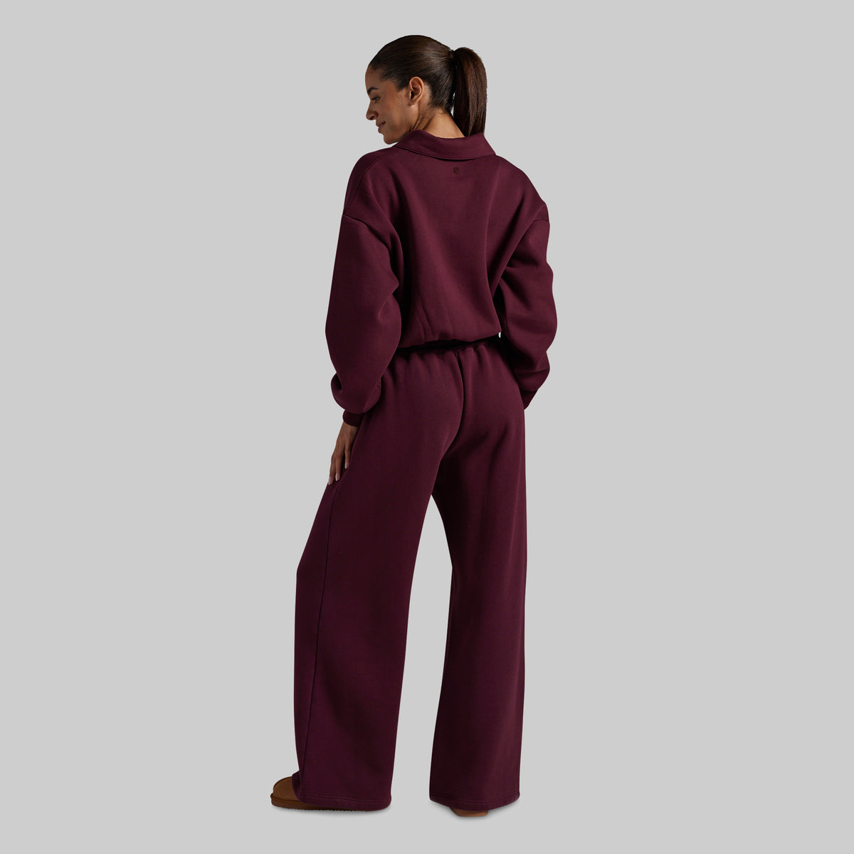 A model shown from the back wearing a matching deep burgundy two-piece lounge set consisting of a cropped long-sleeve top and wide-leg pants, set against a solid light gray background.