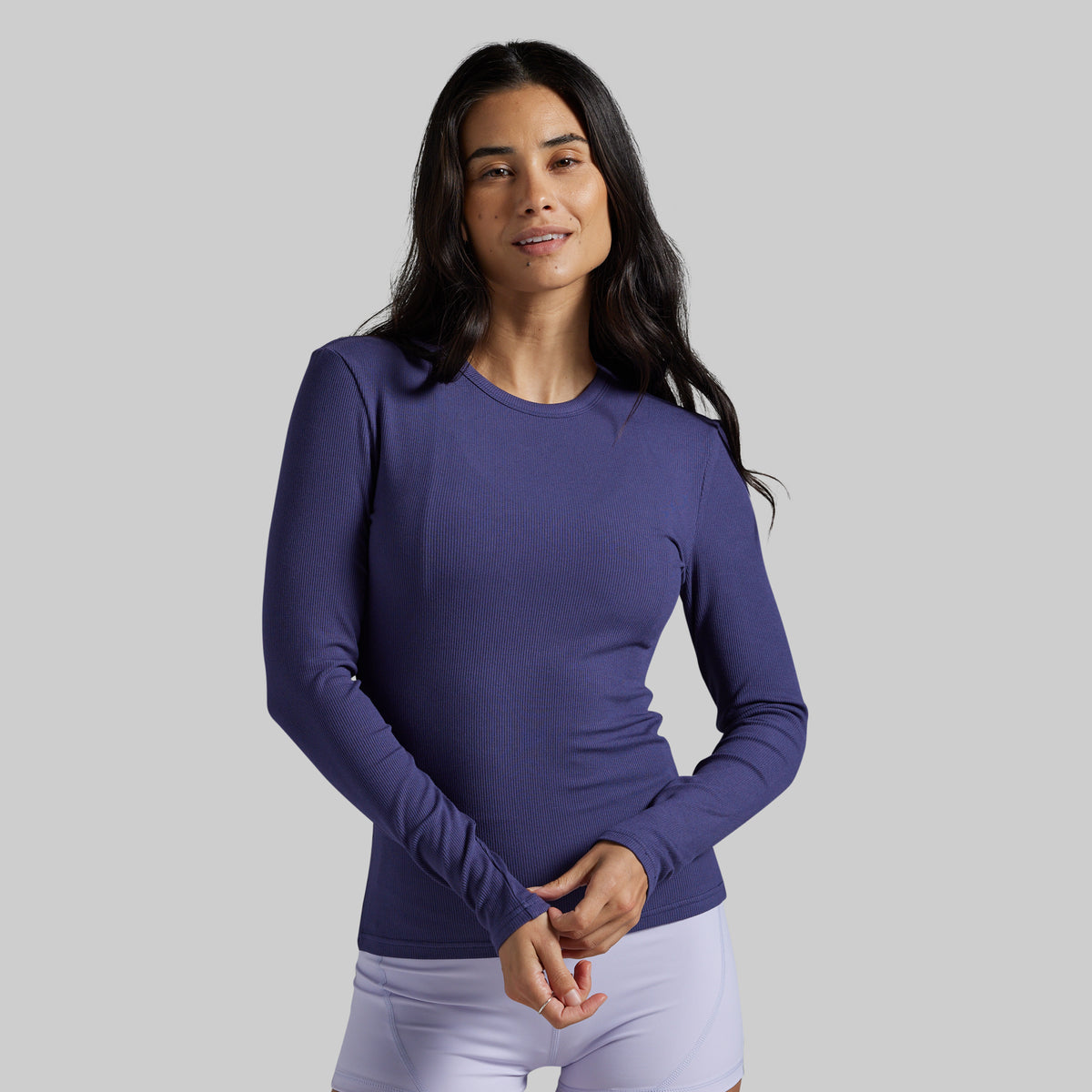 A woman modeling a long-sleeve crewneck athletic top in a solid periwinkle purple color, paired with white leggings, against a solid light gray background.