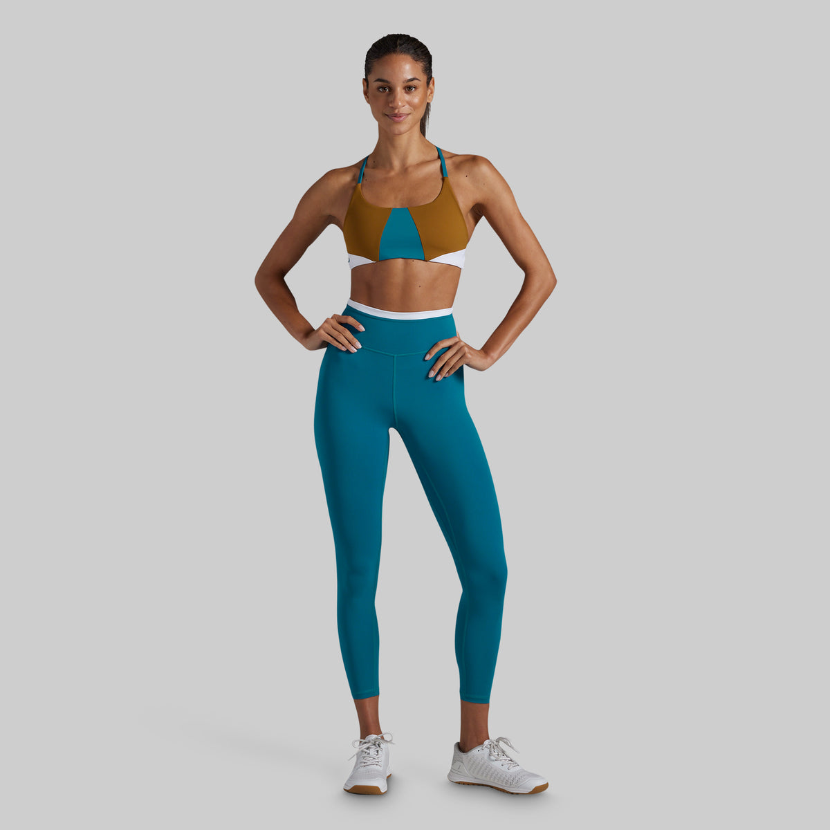 A woman modeling a two-piece activewear set consisting of a color-blocked sports bra in brown, teal, and white, paired with high-waisted teal leggings, against a solid gray background.