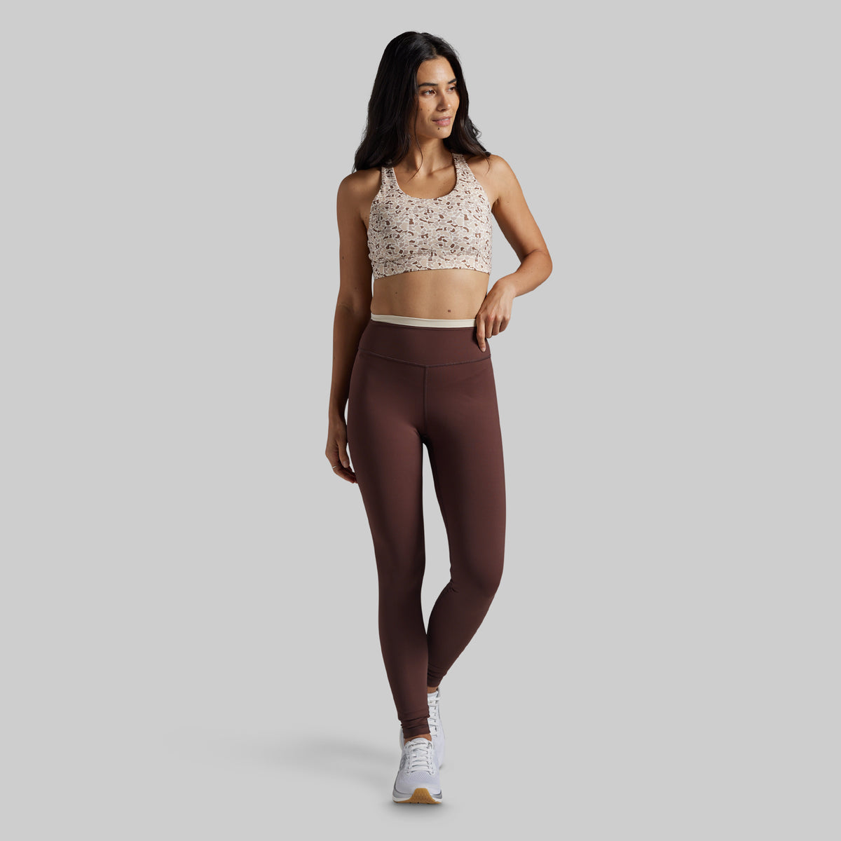 A woman modeling a matching activewear set consisting of a beige and brown patterned sports bra and high-waisted dark brown leggings, standing against a solid gray studio background.