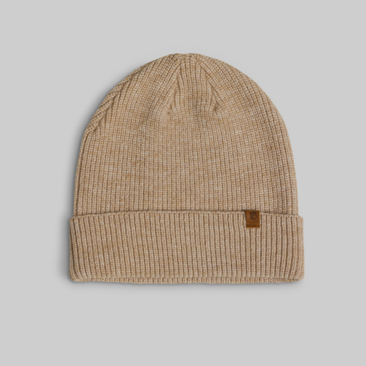 A beige ribbed knit beanie hat with a small brown leather brand patch on the folded cuff, shown against a solid gray background.