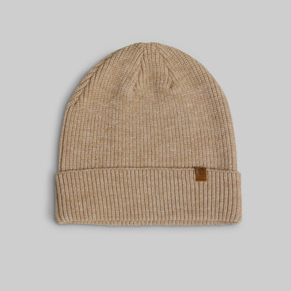 A beige ribbed knit beanie hat with a small brown leather brand patch on the folded cuff, shown against a solid gray background.