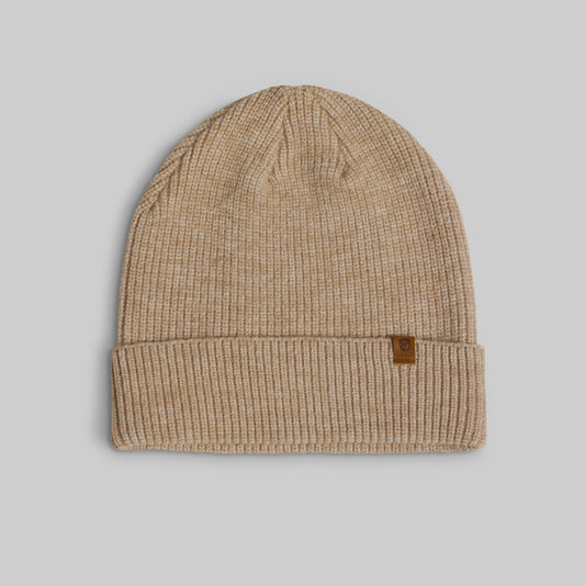 A beige ribbed knit beanie hat with a small brown leather brand patch on the folded cuff, shown against a solid gray background.