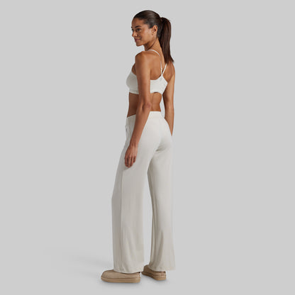 A woman stands in profile wearing a matching white two-piece lounge set consisting of a lounge bra and wide-leg pants, paired with beige slippers, against a solid light gray background.