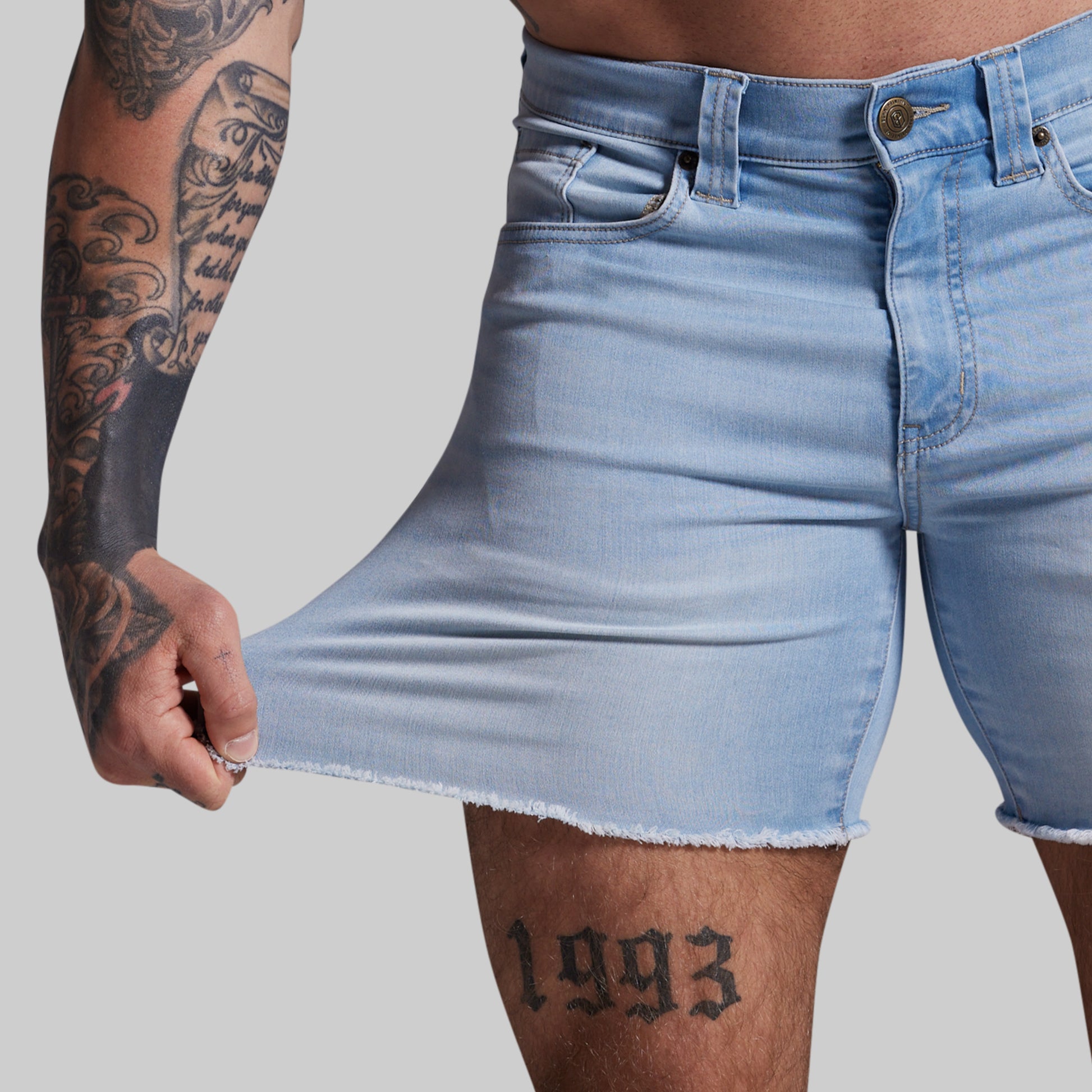 Person wearing light blue denim shorts with tattoos on arm and leg against a gray background