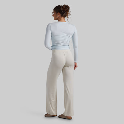A woman shown from the back wearing a light blue long-sleeve top and cream wide-leg pants, standing against a plain light gray studio background.