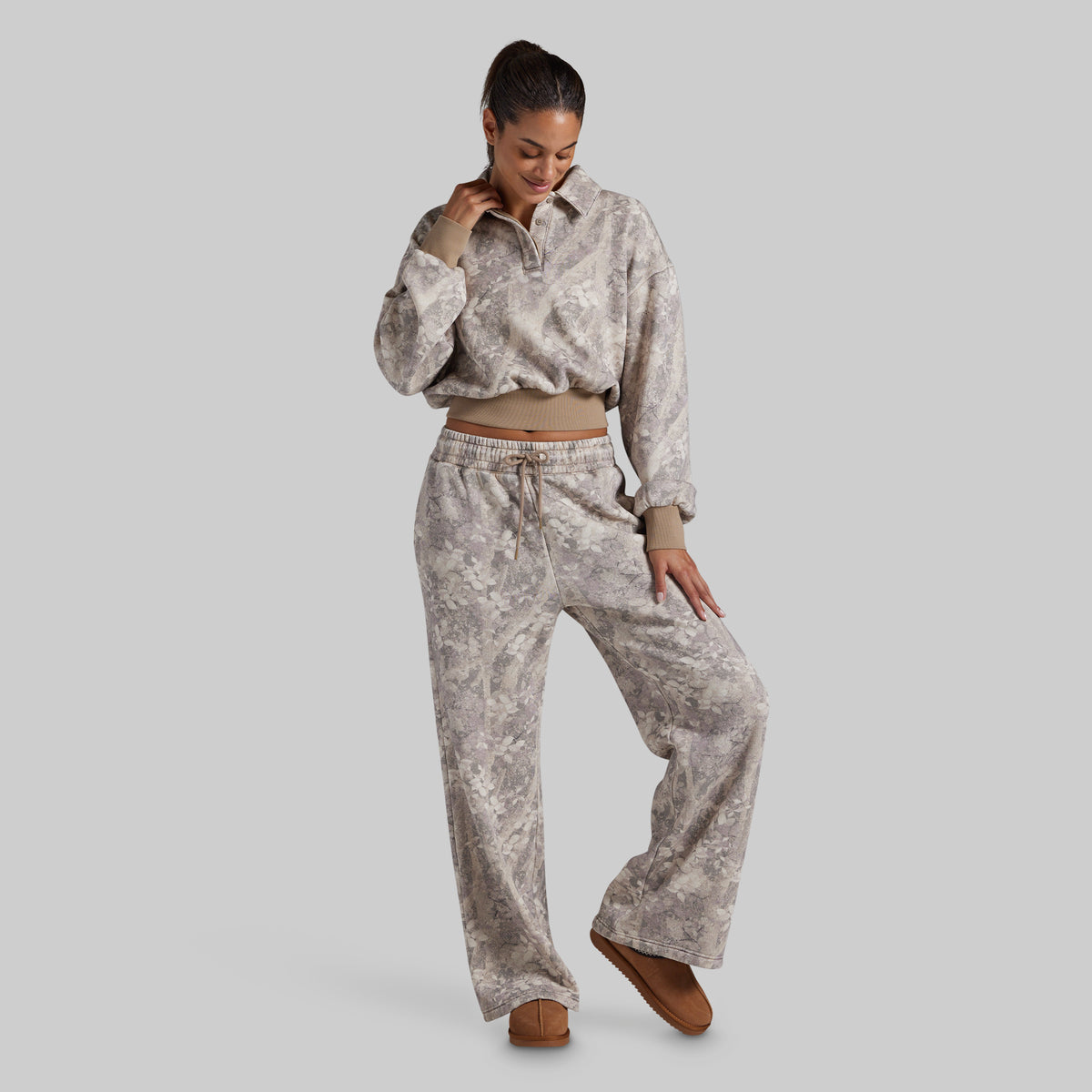 A woman modeling a matching two-piece loungewear set featuring a long-sleeve cropped polo sweatshirt and wide-leg sweatpants in a light gray and beige abstract pattern, styled with brown suede shoes against a solid gray background.