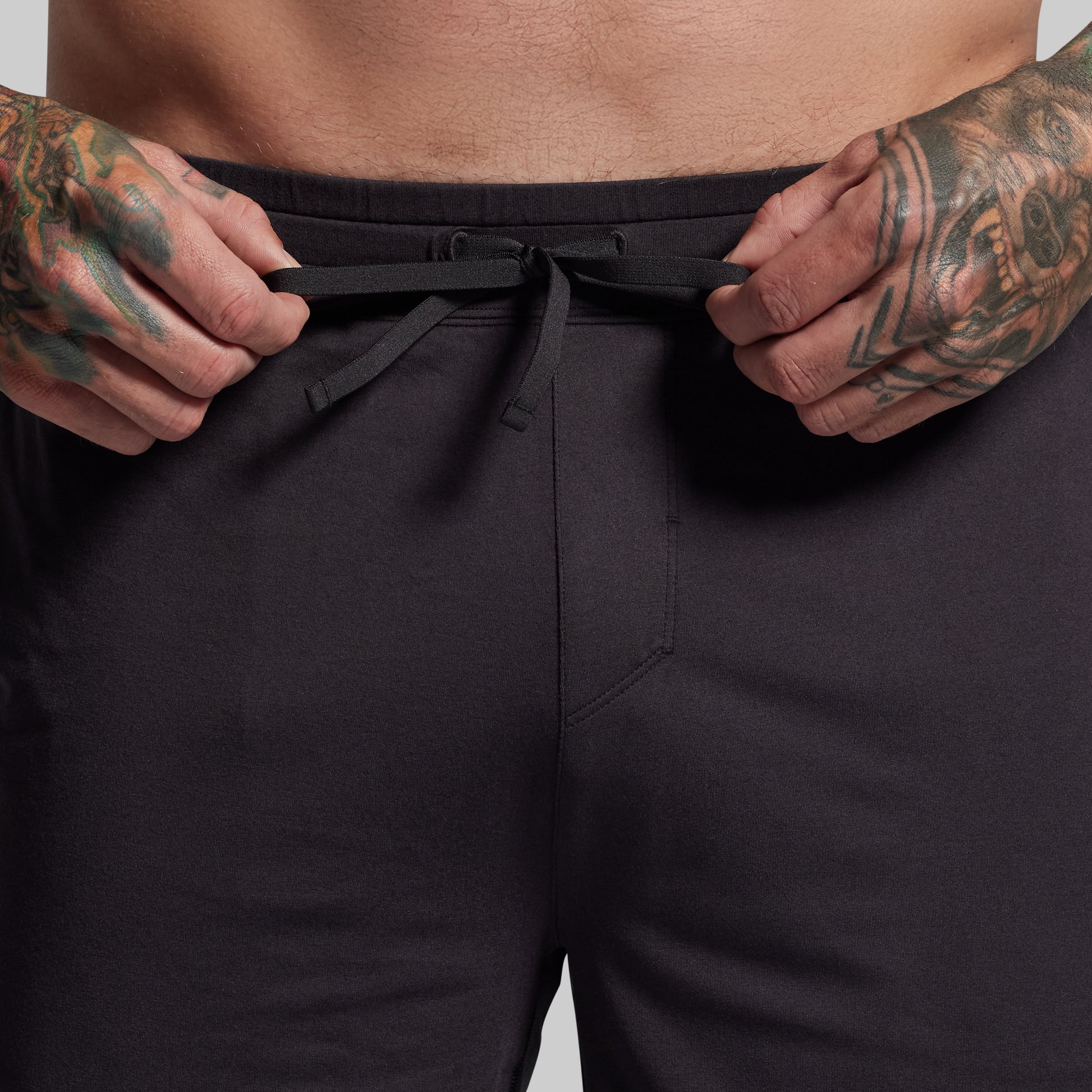 Person wearing black shorts with tattooed hands adjusting the waistband against a neutral background