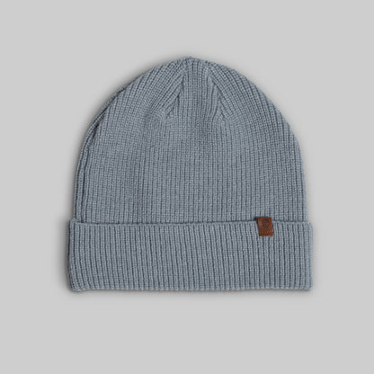 A light blue-gray ribbed knit beanie hat with a small brown leather brand tab on the cuff, shown against a solid gray background.
