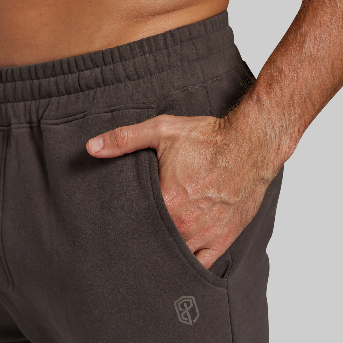 Close-up of a person wearing dark grey athletic sweatpants, showing the side pocket with a hand inside and a small geometric logo near the lower leg on a light gray background.