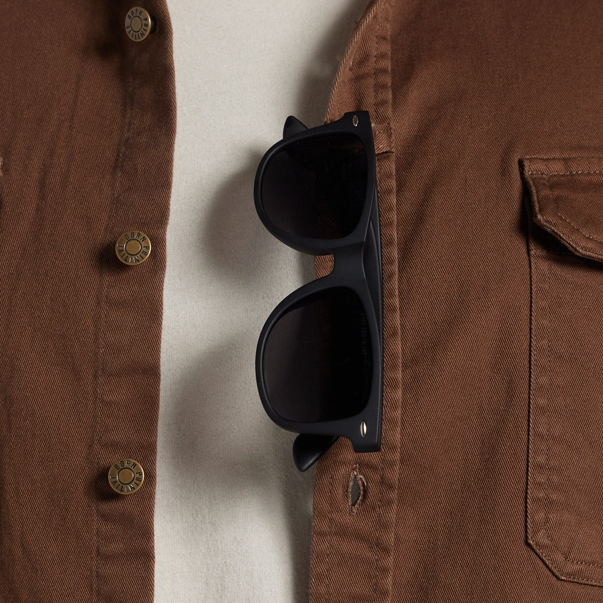 Black sunglasses with dark lenses hanging from a brown button-up shirt pocket, shown in a close-up lifestyle shot.