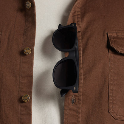 Black sunglasses with dark lenses hanging from a brown button-up shirt pocket, shown in a close-up lifestyle shot.