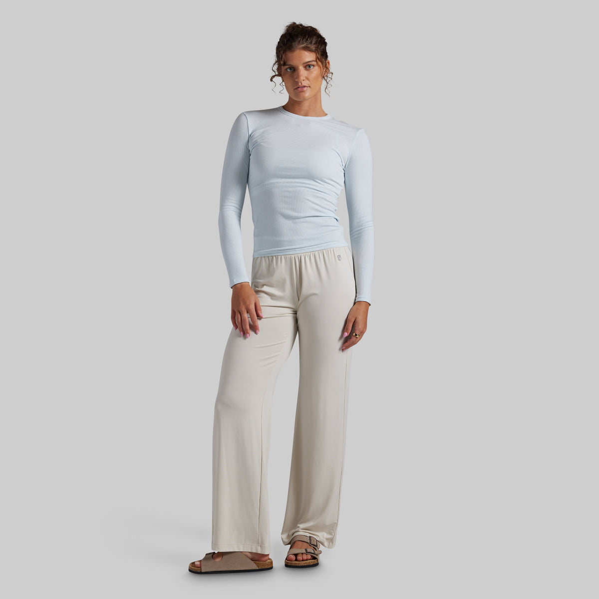 A woman modeling a light blue long-sleeve top and cream wide-leg pants against a solid light gray studio background.