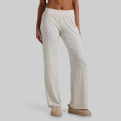 A model wearing a pair of off-white wide-leg lounge pants and beige slip-on shoes, shown from the waist down against a solid gray background.