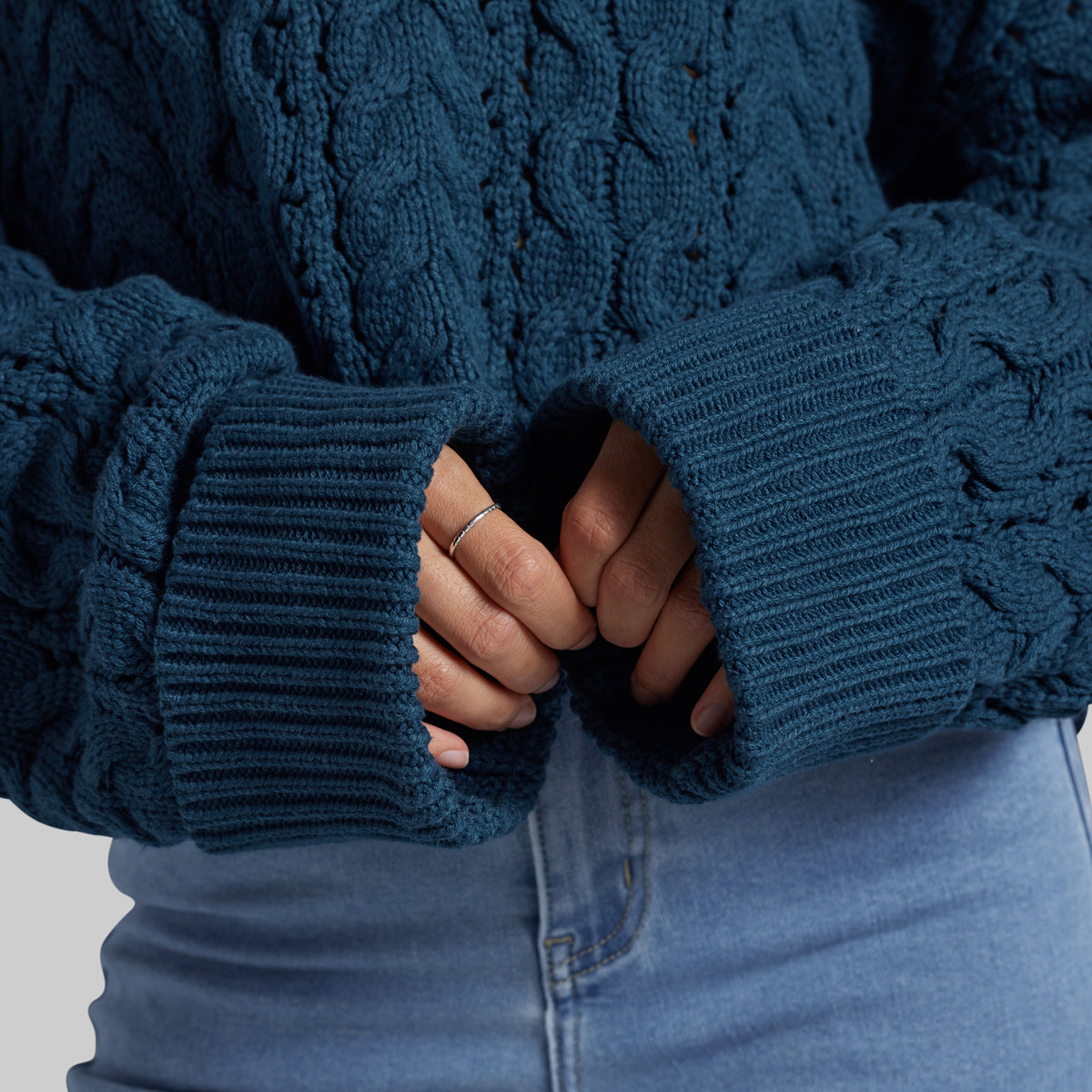 Close-up of a person wearing a dark teal cable-knit sweater with ribbed cuffs, paired with light blue jeans. The person's hands are tucked inside the cuffs.
