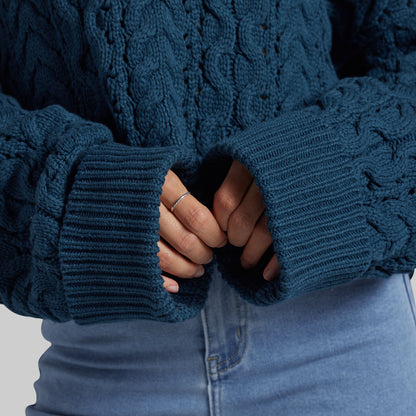 Close-up of a person wearing a dark teal cable-knit sweater with ribbed cuffs, paired with light blue jeans. The person's hands are tucked inside the cuffs.