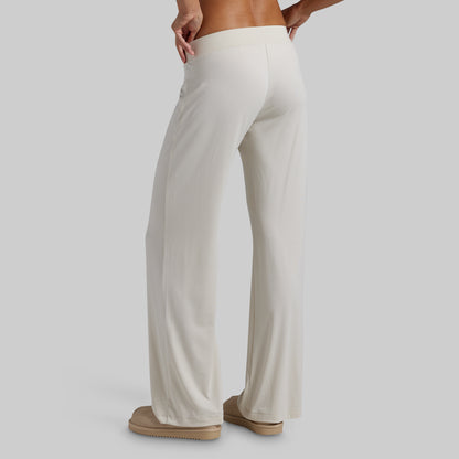 A person shown from the back wearing loose, wide-leg pants in a warm off-white or cream color, paired with tan slip-on shoes, against a plain light gray studio background.