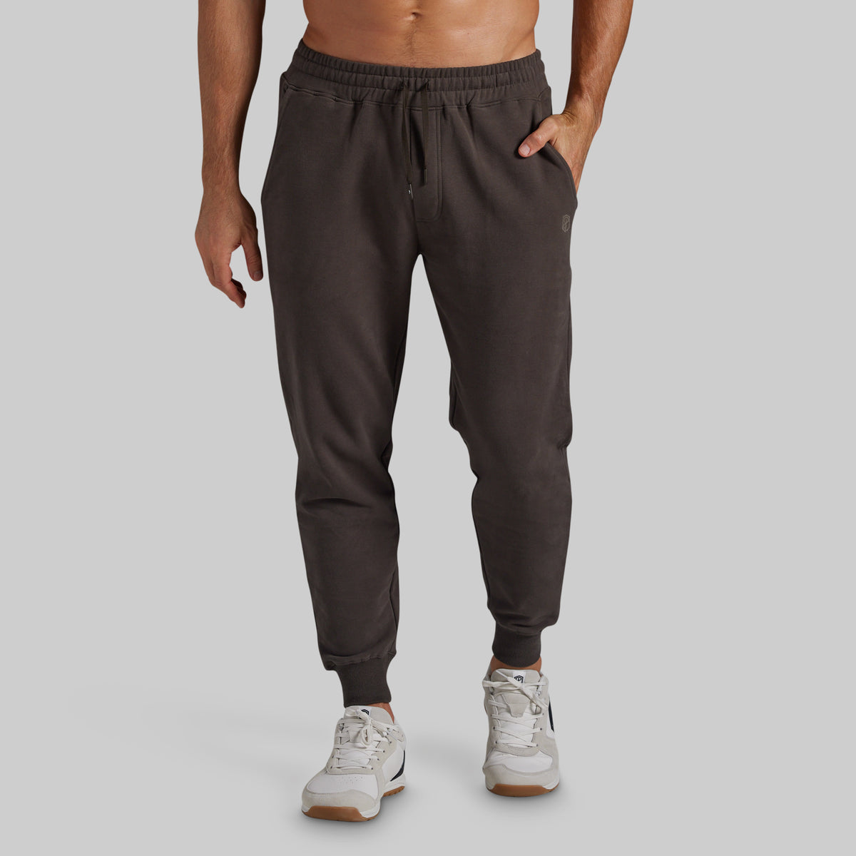 A man wearing dark brown jogger sweatpants with an elastic drawstring waist and ribbed ankle cuffs, paired with white sneakers, shown from the waist down against a solid gray background.