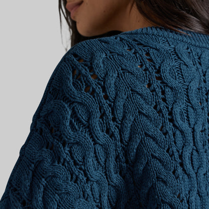 Close-up of a person wearing a textured cable-knit sweater in a deep teal blue color, showing the intricate knit pattern and shoulder area against a neutral light gray background.