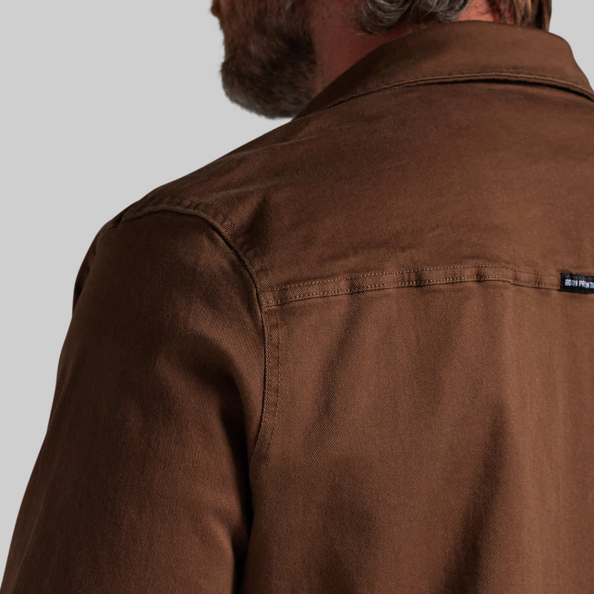 Close-up back view of a man wearing a brown overshirt or jacket, showing the collar, shoulder, and a small black brand tab on the upper right shoulder against a light gray background.