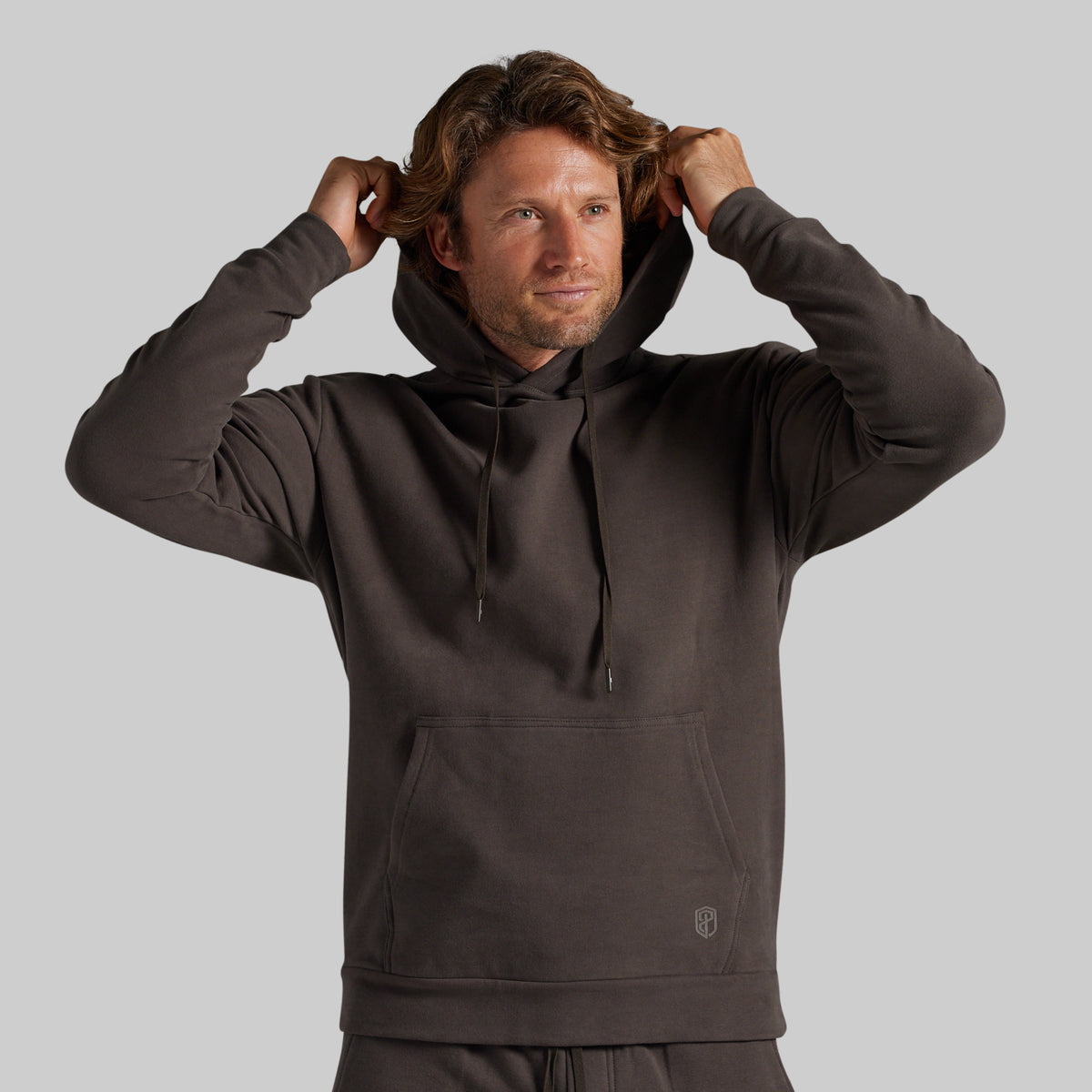 A man wearing a dark brown pullover hoodie with drawstrings and a kangaroo pocket, adjusting his hood against a solid gray background.