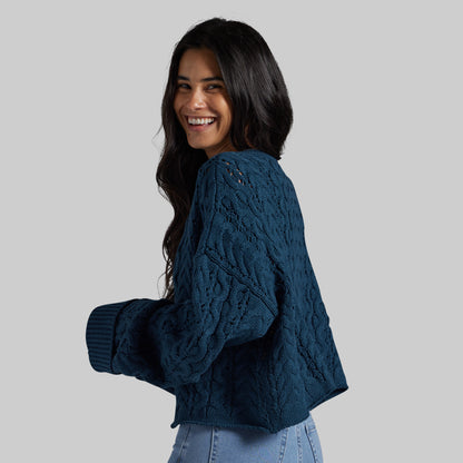 A woman with long dark hair is shown from the back and side, wearing a textured navy blue cable-knit cropped sweater with ribbed cuffs, paired with light blue jeans, against a solid light gray background.