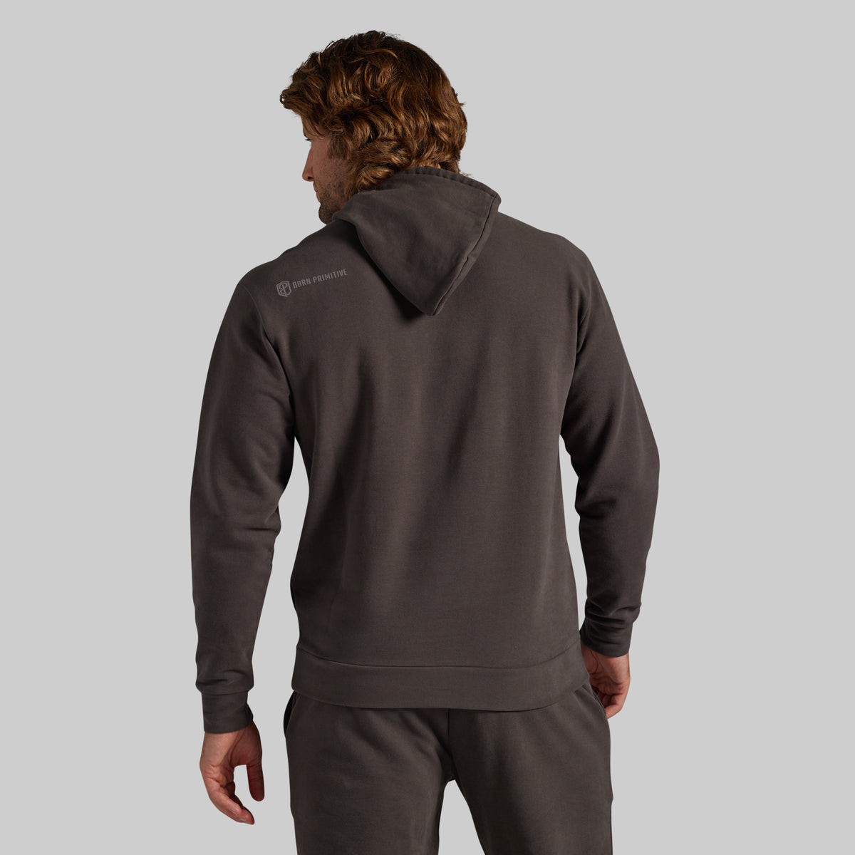 A man shown from the back wearing a dark charcoal gray hoodie and matching sweatpants against a solid light gray background.