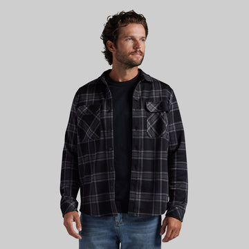 A man wearing a black and gray plaid button-up flannel shirt over a black t-shirt and blue jeans, standing against a solid gray background.