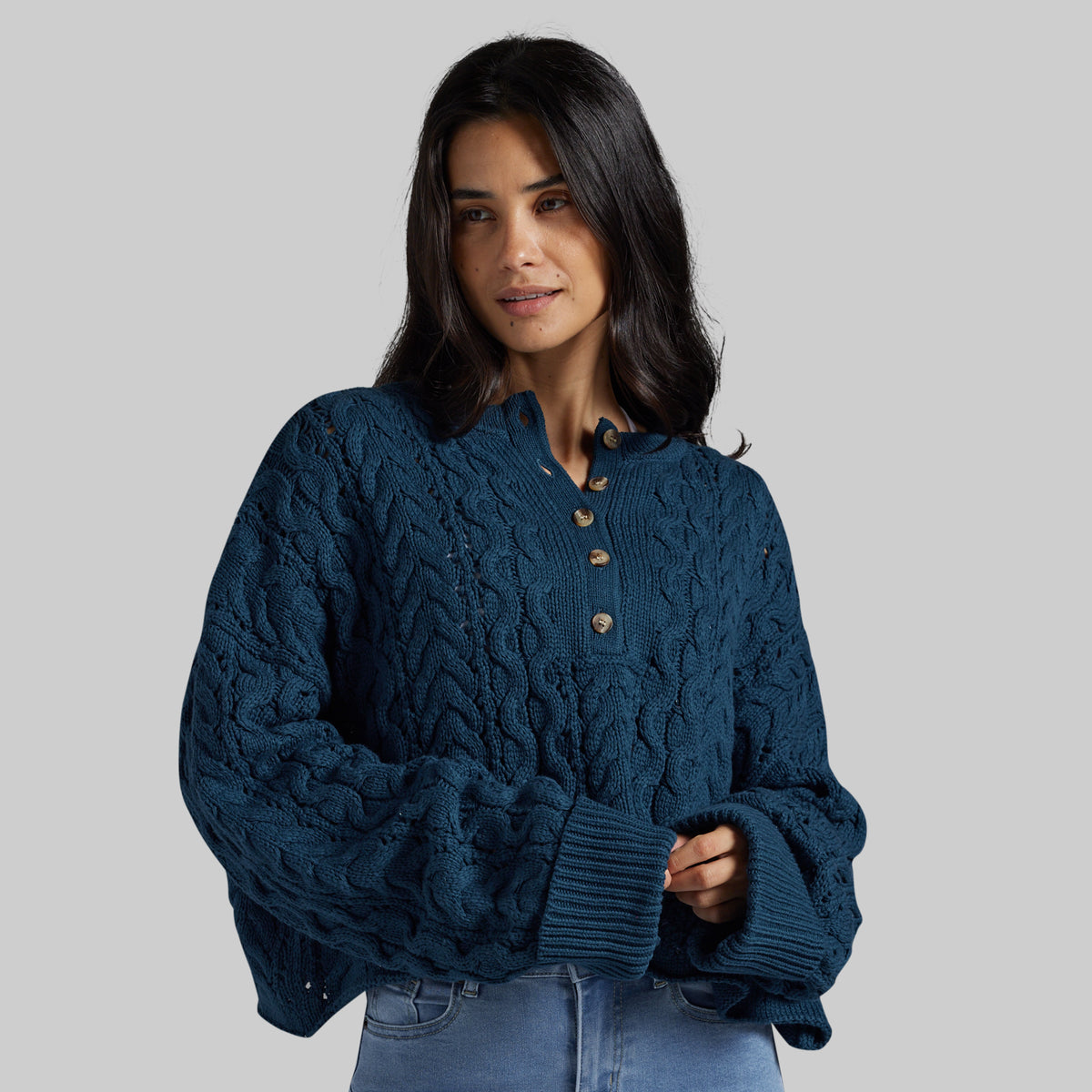 A woman wearing a dark teal cable-knit cropped sweater with a buttoned henley neckline, styled with light blue jeans against a solid gray studio background.