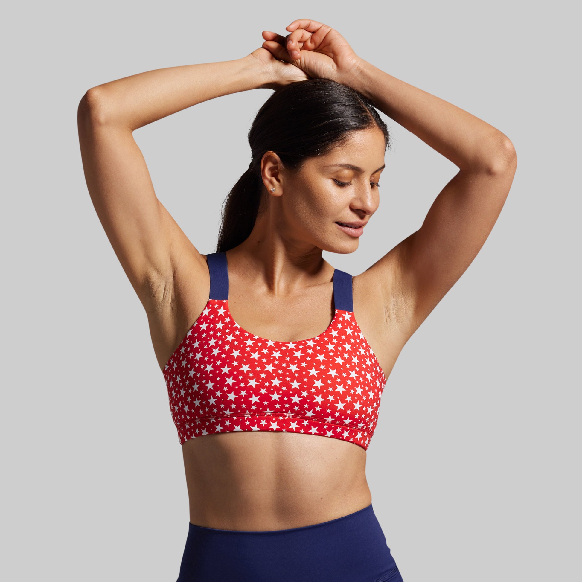 Woman wearing a red sports bra with white star pattern and blue leggings on a gray background