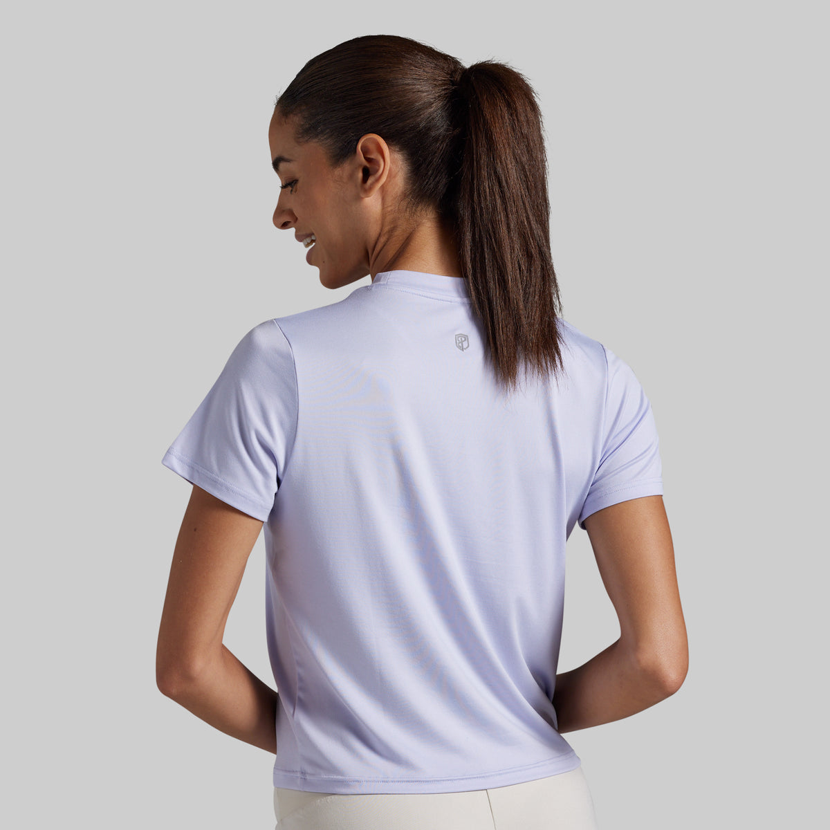 A woman with her hair in a ponytail is shown from the back wearing a light lavender short-sleeve athletic t-shirt. 