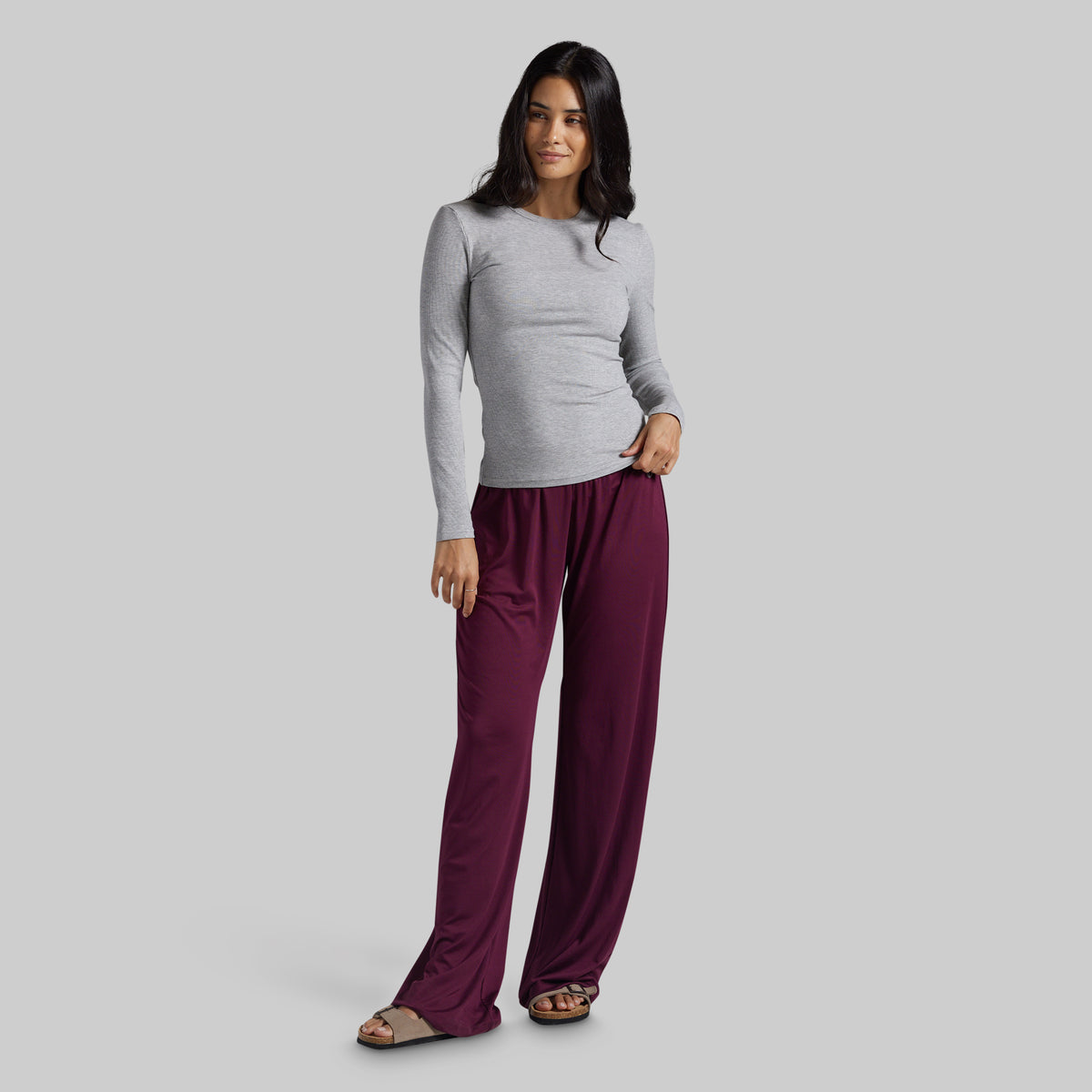 A woman modeling a light gray long-sleeve top and burgundy wide-leg pants against a solid gray studio background.