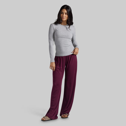 A woman modeling a light gray long-sleeve top and burgundy wide-leg pants against a solid gray studio background.