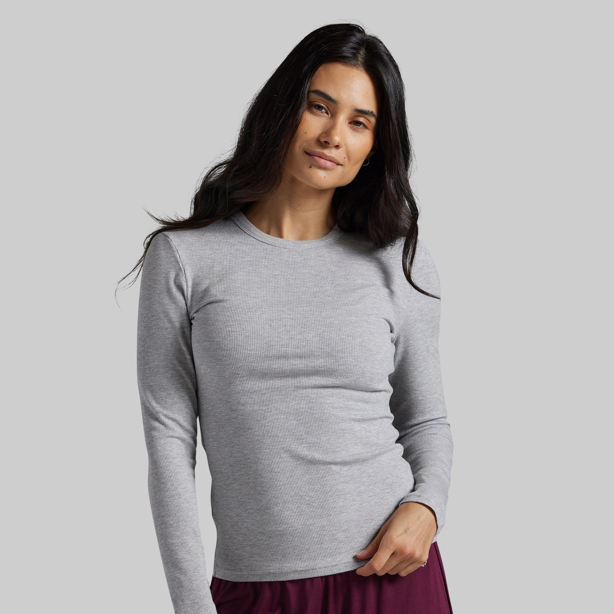 A woman modeling a light gray long-sleeve crewneck top against a solid gray background.