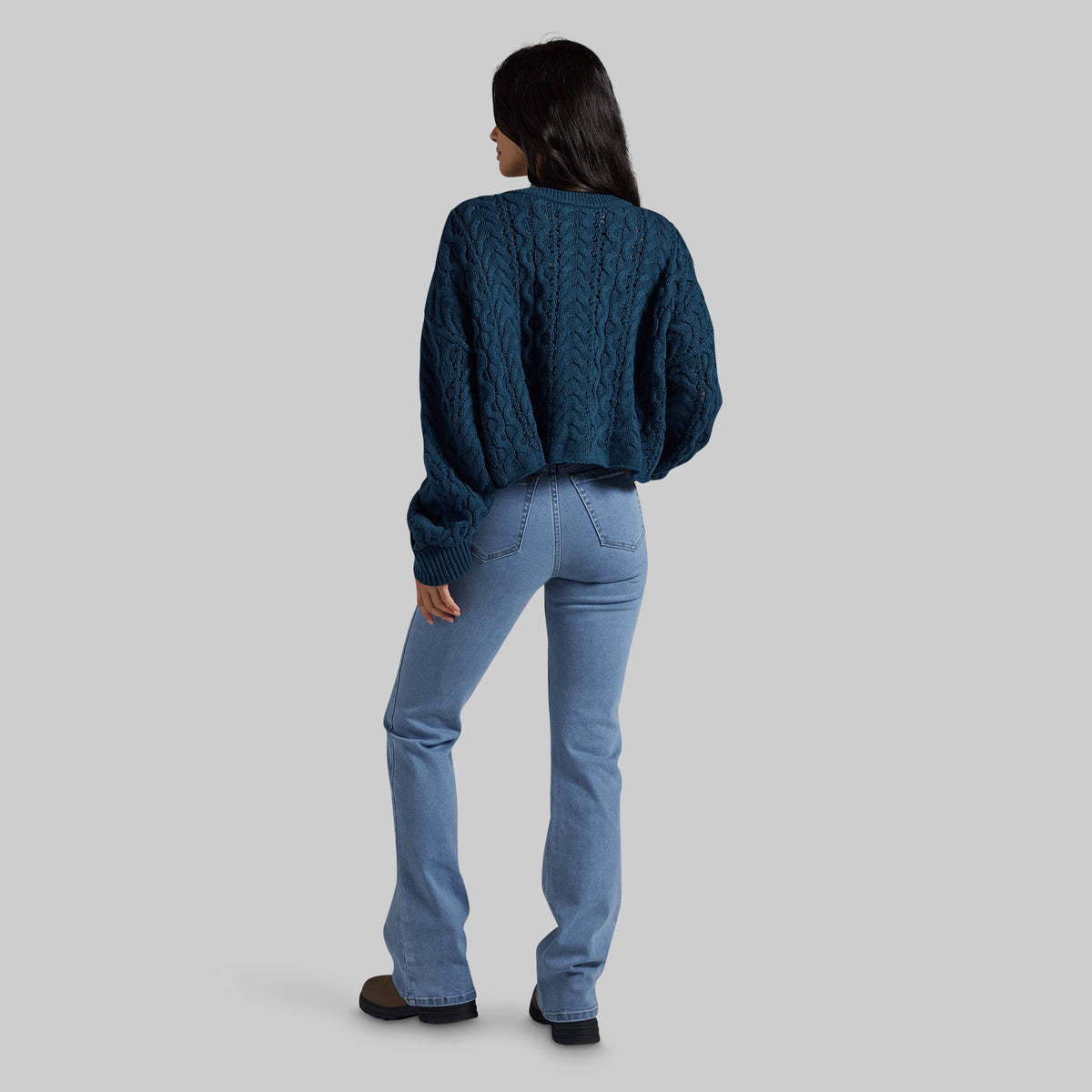 A woman shown from the back wearing a dark teal cable-knit cropped sweater and light blue jeans, standing against a solid gray studio background.