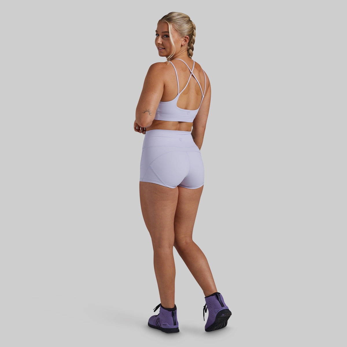 A woman modeling a light purple/lavender activewear set consisting of a sports bra and high-waisted shorts, shown from a rear three-quarter view against a solid gray studio background.