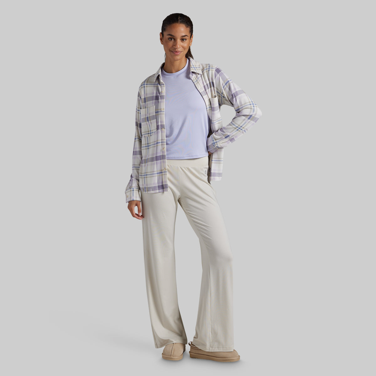 A woman modeling a light purple plaid button-down shirt layered over a light lavender crewneck top, paired with cream-colored wide-leg pants and beige slip-on shoes, against a solid gray studio background.