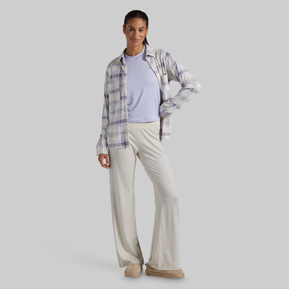 A woman modeling a light purple plaid button-down shirt layered over a light lavender crewneck top, paired with cream-colored wide-leg pants and beige slip-on shoes, against a solid gray studio background.