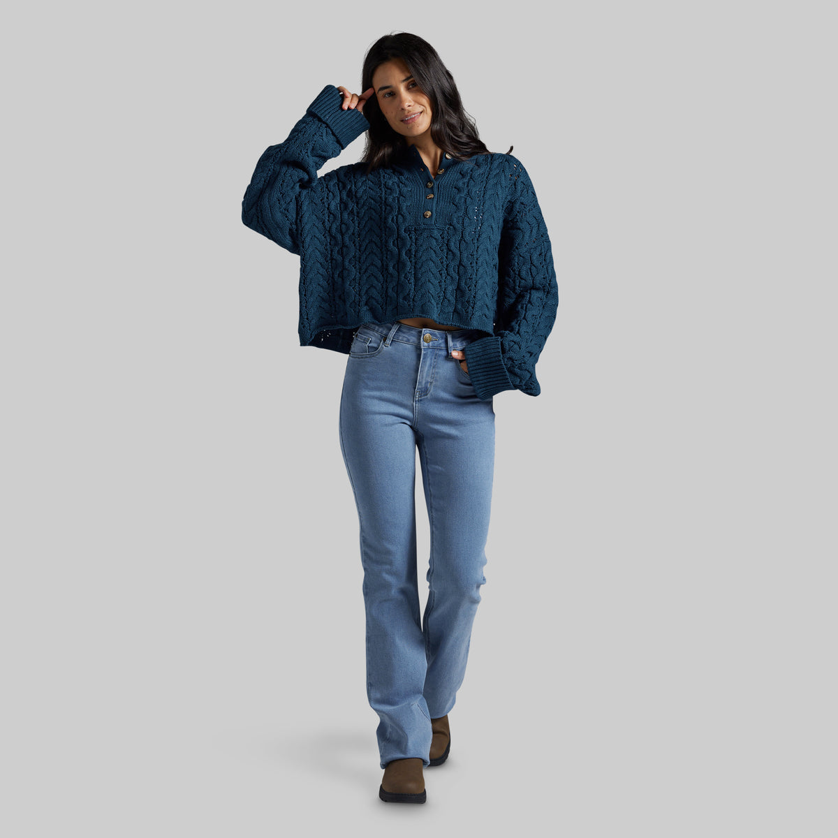 A woman modeling a dark teal blue cable-knit cropped henley sweater with light-wash blue jeans and brown boots, standing against a solid gray studio background.