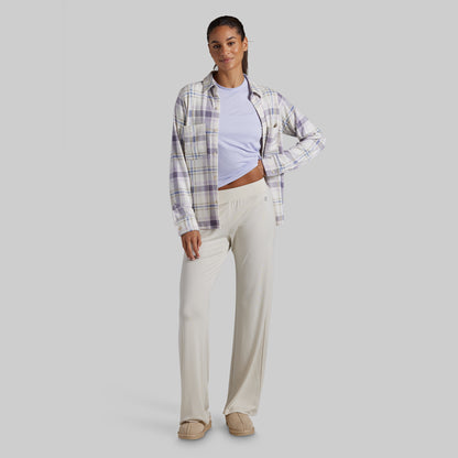A woman modeling a casual outfit consisting of a light purple and white plaid button-up shirt over a light lavender crop top, paired with cream-colored wide-leg pants and beige slip-on shoes, set against a solid gray background.