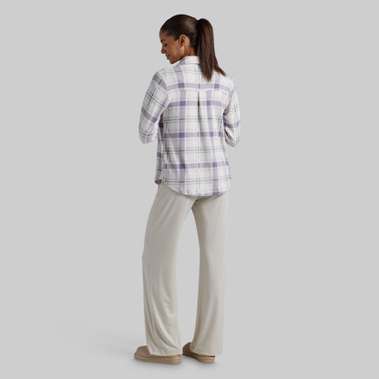 A woman is shown from the back wearing a light purple and white plaid button-down shirt with beige wide-leg pants and tan slippers, set against a solid light gray background.