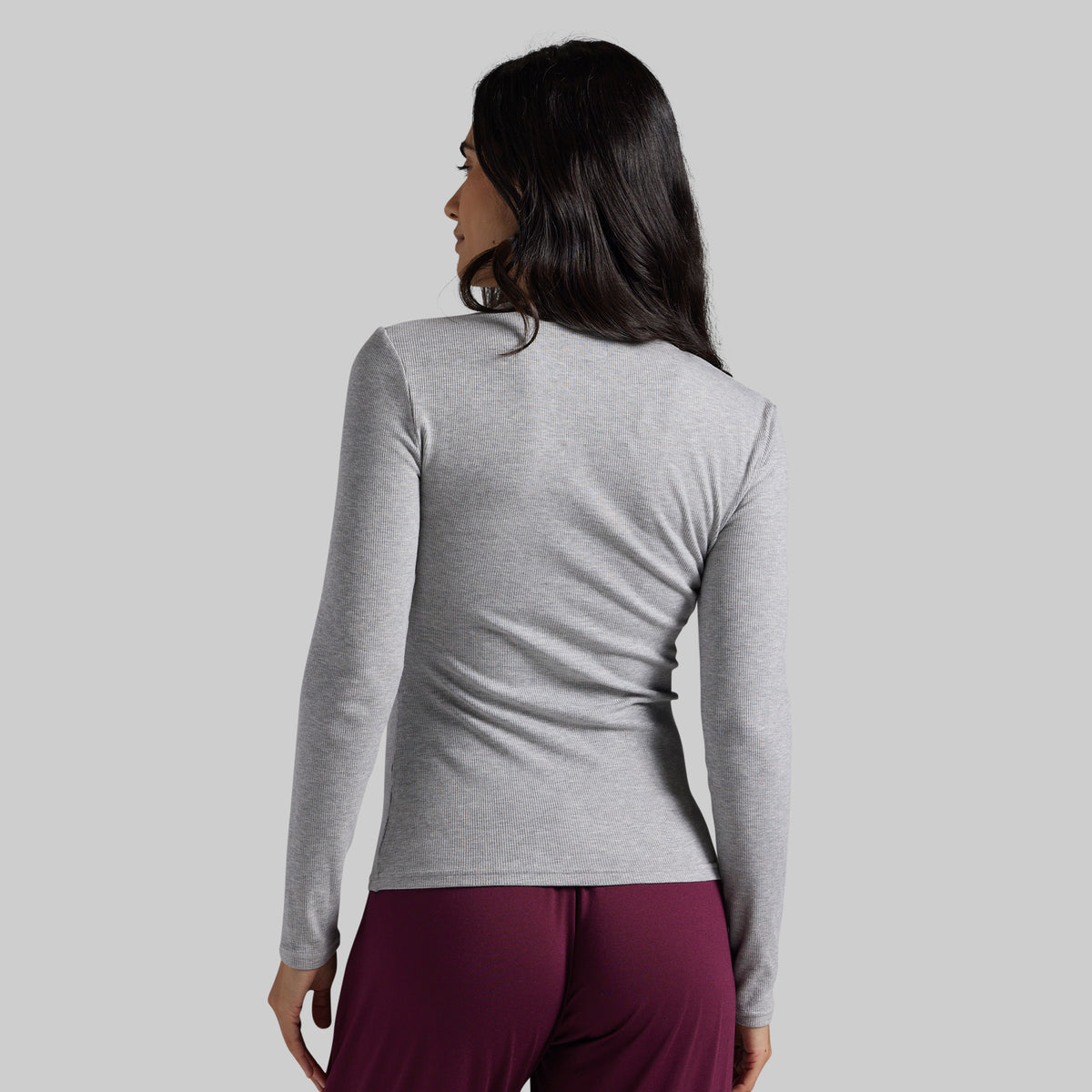 A woman shown from the back wearing a light heather gray long-sleeve athletic top and dark burgundy pants against a solid light gray background.