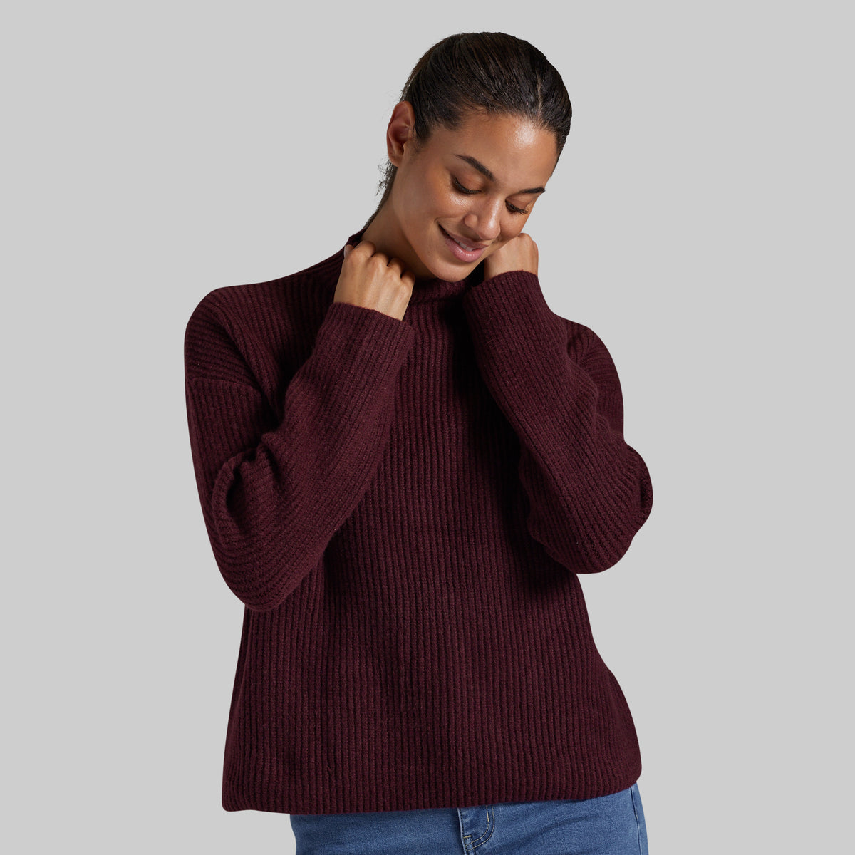 A woman wearing a ribbed knit mock neck sweater in deep burgundy, paired with blue jeans, against a solid gray studio background.