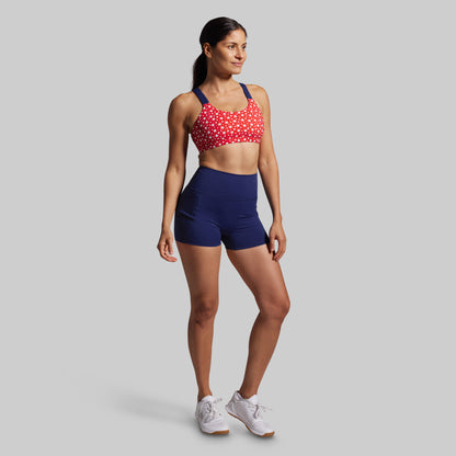Woman wearing a red sports bra with white patterns and blue shorts on a gray background