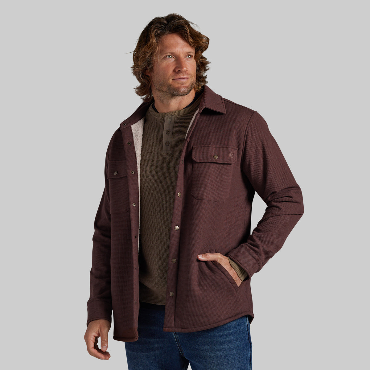 A man wearing a dark brown button-up jacket with a light beige sherpa lining over a brown henley shirt and blue jeans, photographed against a solid gray background.