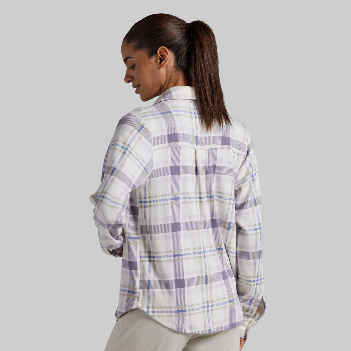 A woman shown from the back wearing a long-sleeve plaid button-up shirt in white, lavender, and light blue, paired with light beige pants, against a solid gray background.