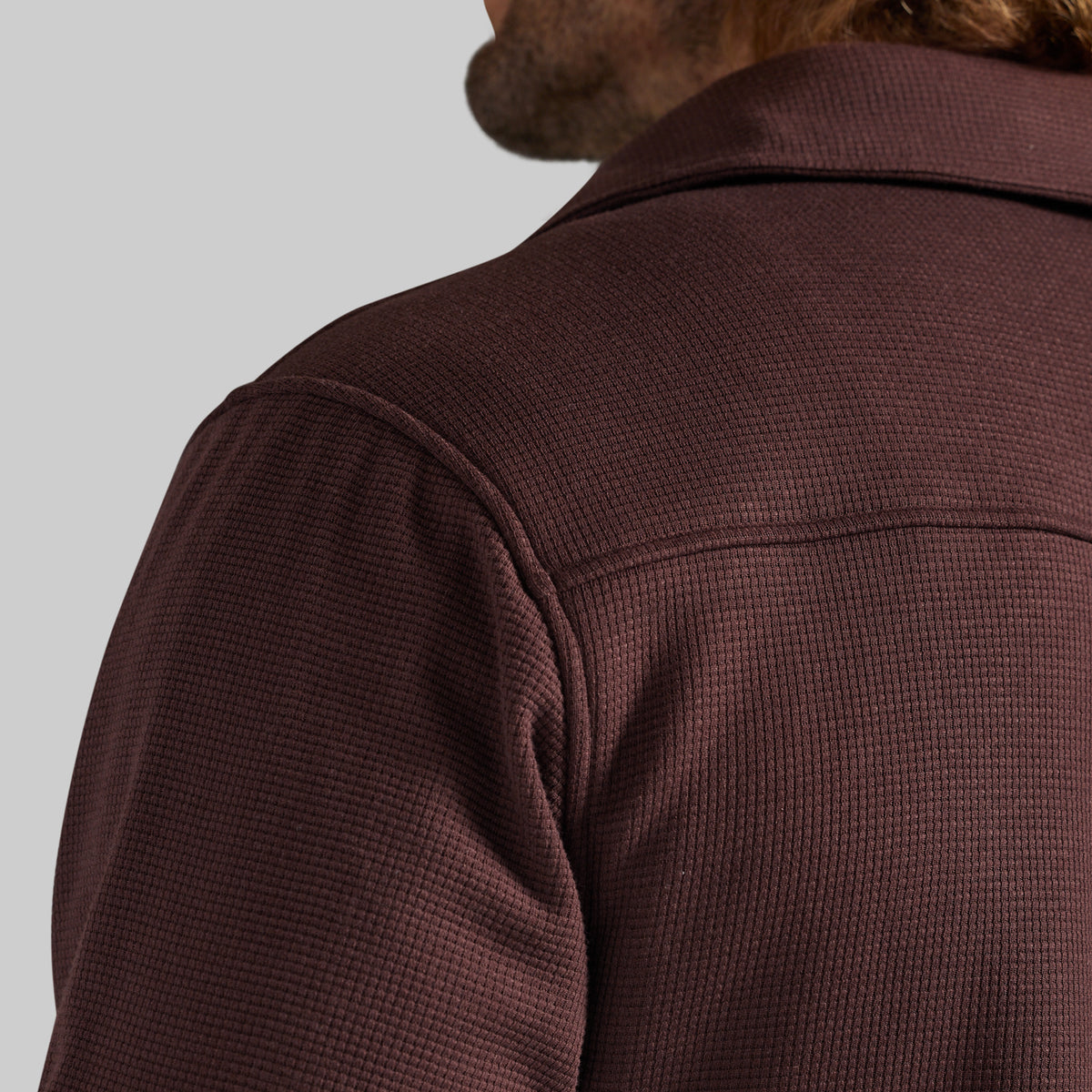 Close-up of a man wearing a textured waffle-knit jacket in a deep burgundy brown, showing the back of the neck and shoulder against a light gray background.